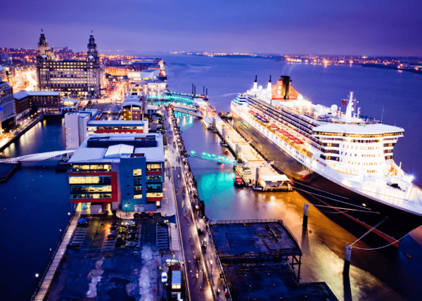 A large Cunard Cruise ship docked at Liverpool cruise terminal at night with the skyline lit up.