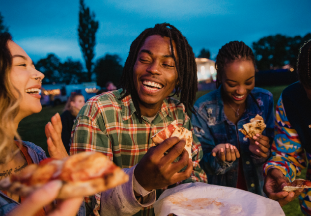 People are smiling eating pizza together in a festival scene.