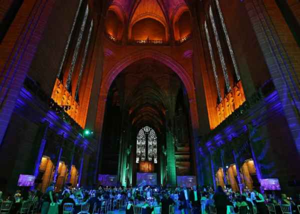 The gothic Liverpool Cathedral vaults in green and blue lights. There are stained glass windows and tables of people.