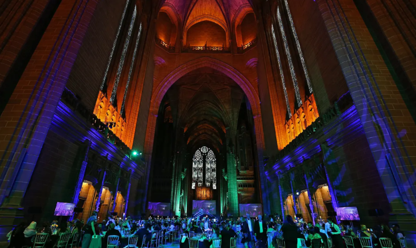 The gothic Liverpool Cathedral vaults in green and blue lights. There are stained glass windows and tables of people.