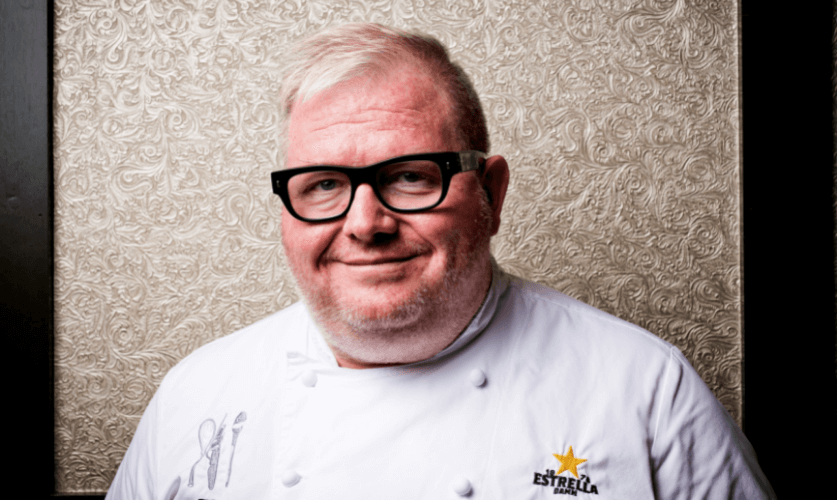 Chef, Paul Skew looks at the camera wearing his chef whites and black glasses.