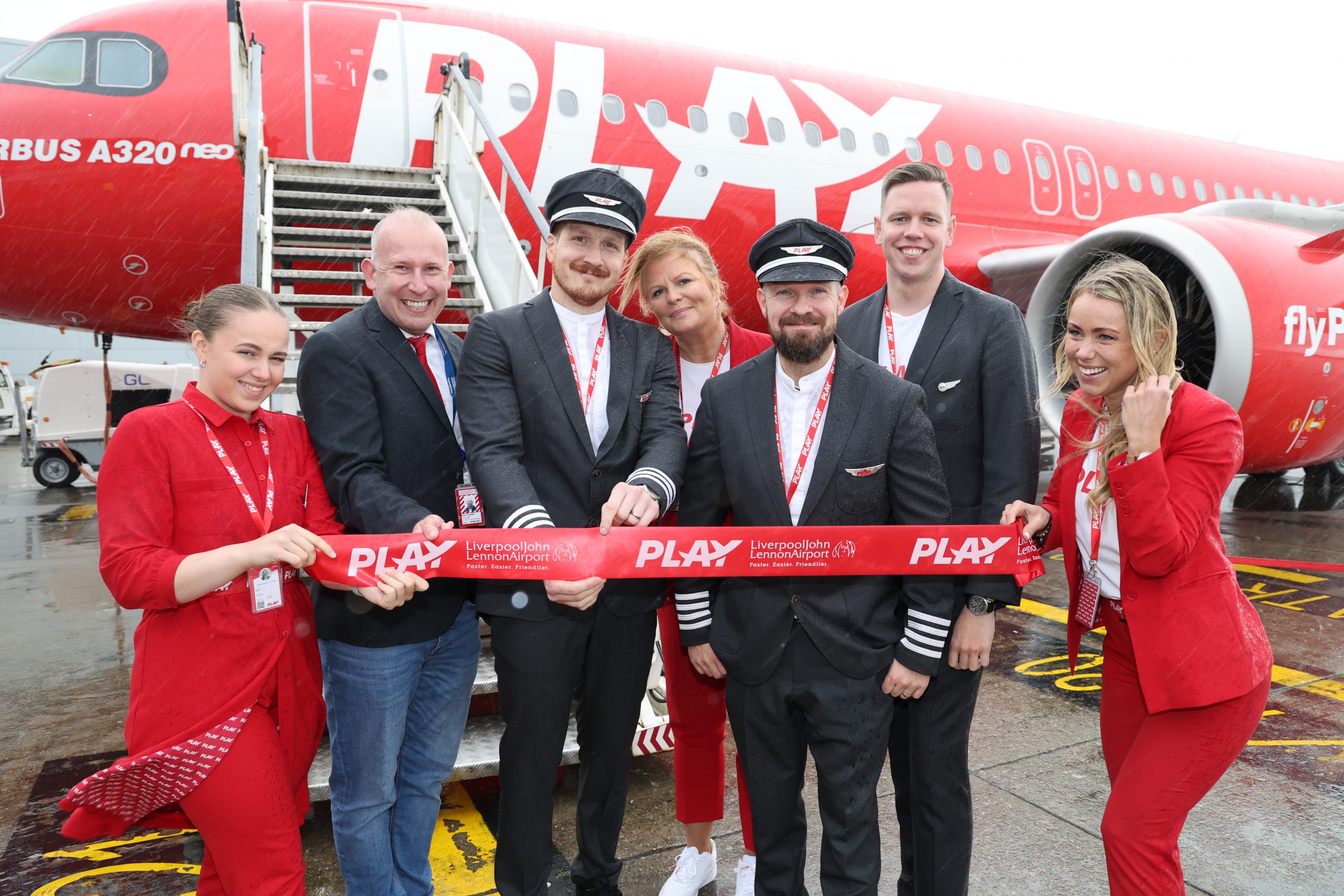 Seven people stood next to a red plane holding up a banner with Play on.