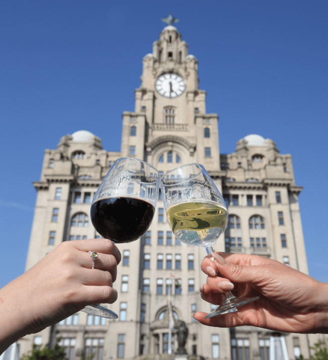 Two wine glasses, one filled with red wine and one filled with white wine are held in front of the Liver building in a 'cheers' position.
